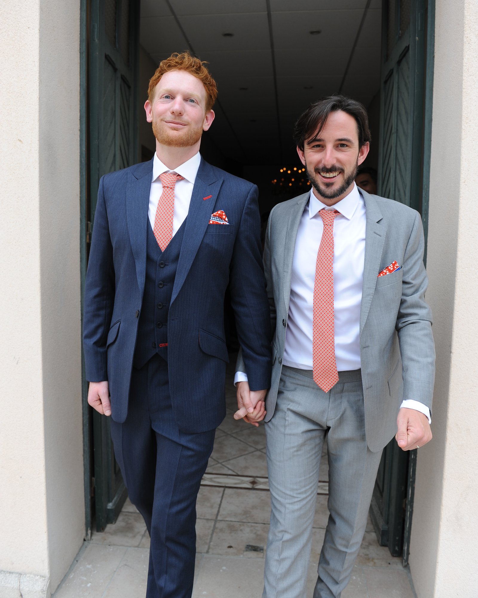 Two grooms hand in hand — a navy three-piece and a light grey suit with matching coral ties