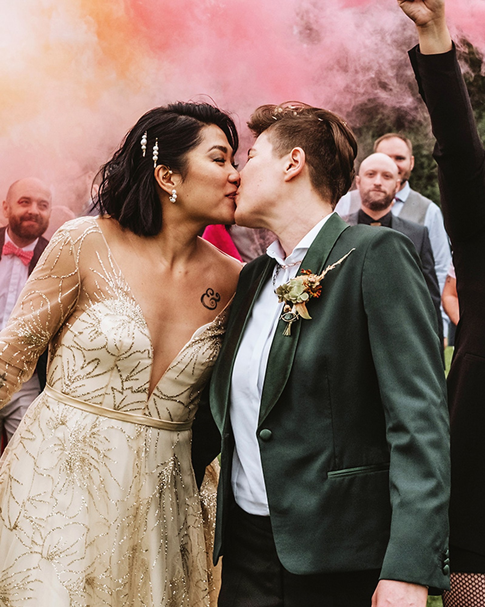 A couple kissing under pink smoke — a gold sequin wedding dress beside a green velvet tuxedo