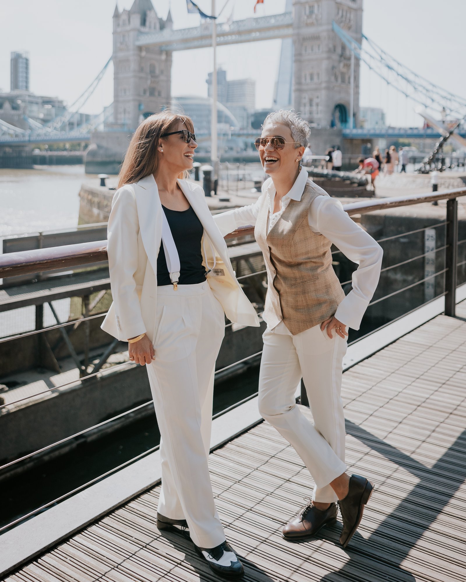 A couple laughing by Tower Bridge — a white trouser suit beside a check waistcoat and white shirt