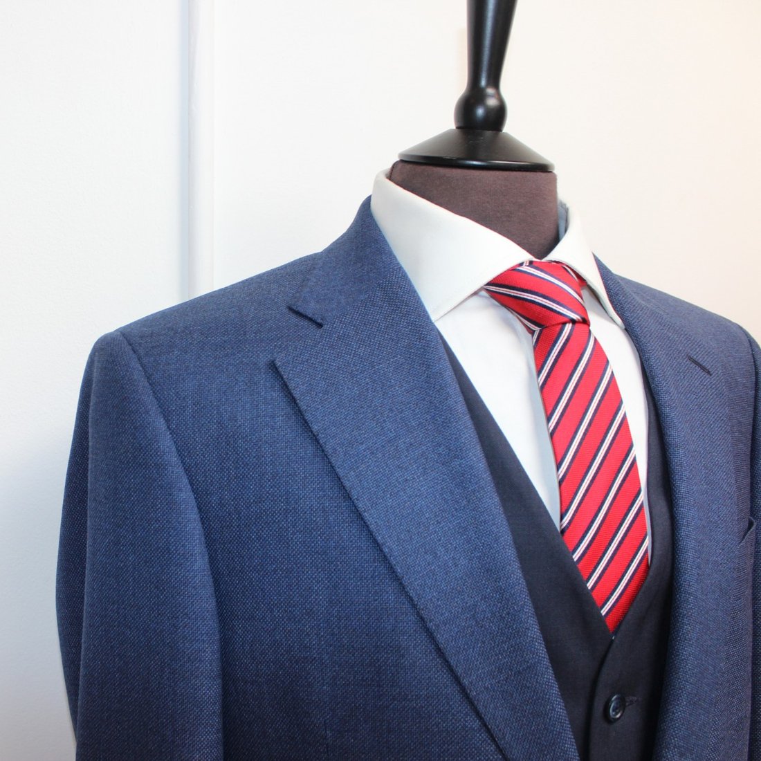 A blue birdseye three-piece suit with a striped red tie on the studio stand
