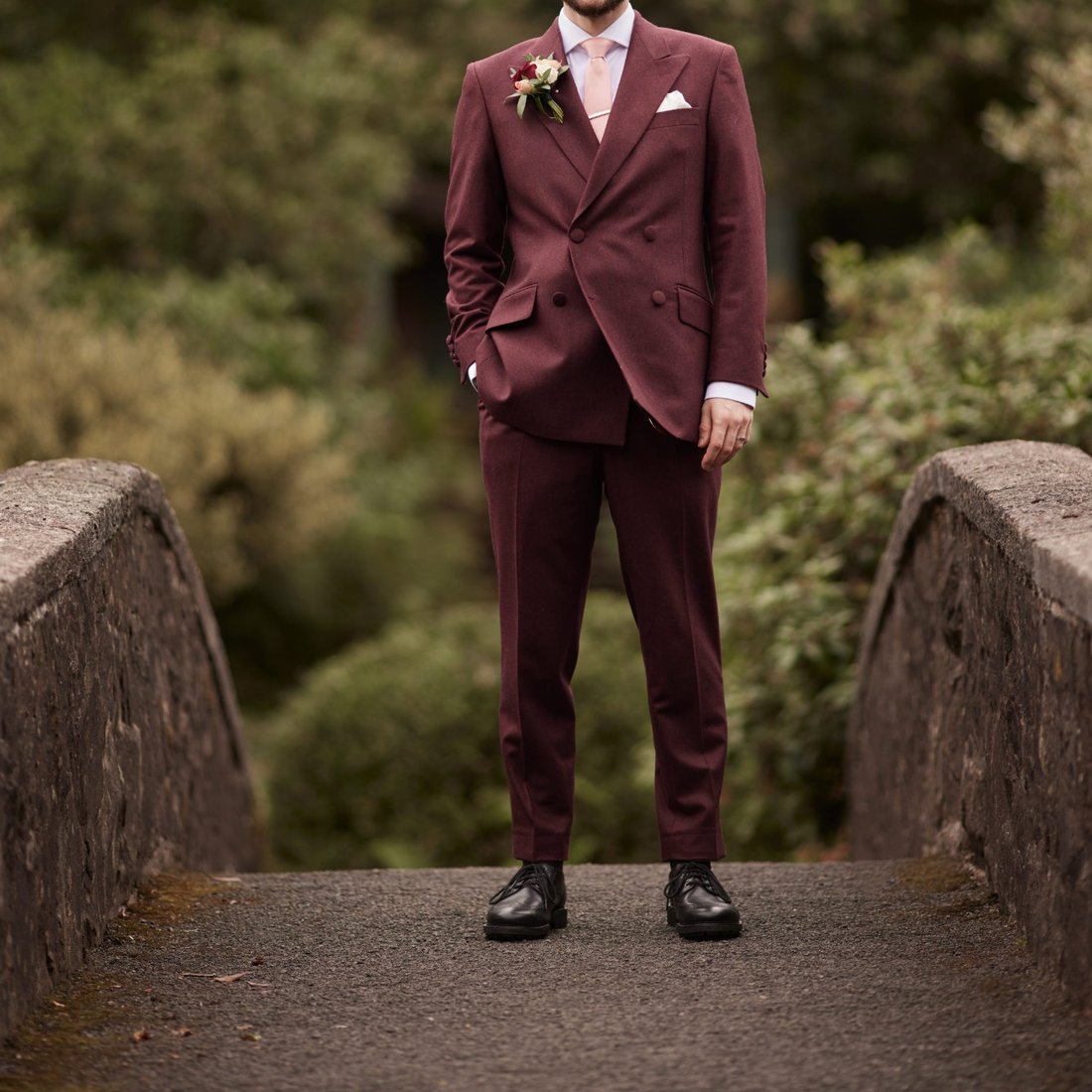 A groom in a double-breasted burgundy wedding suit with blush tie, on a stone bridge