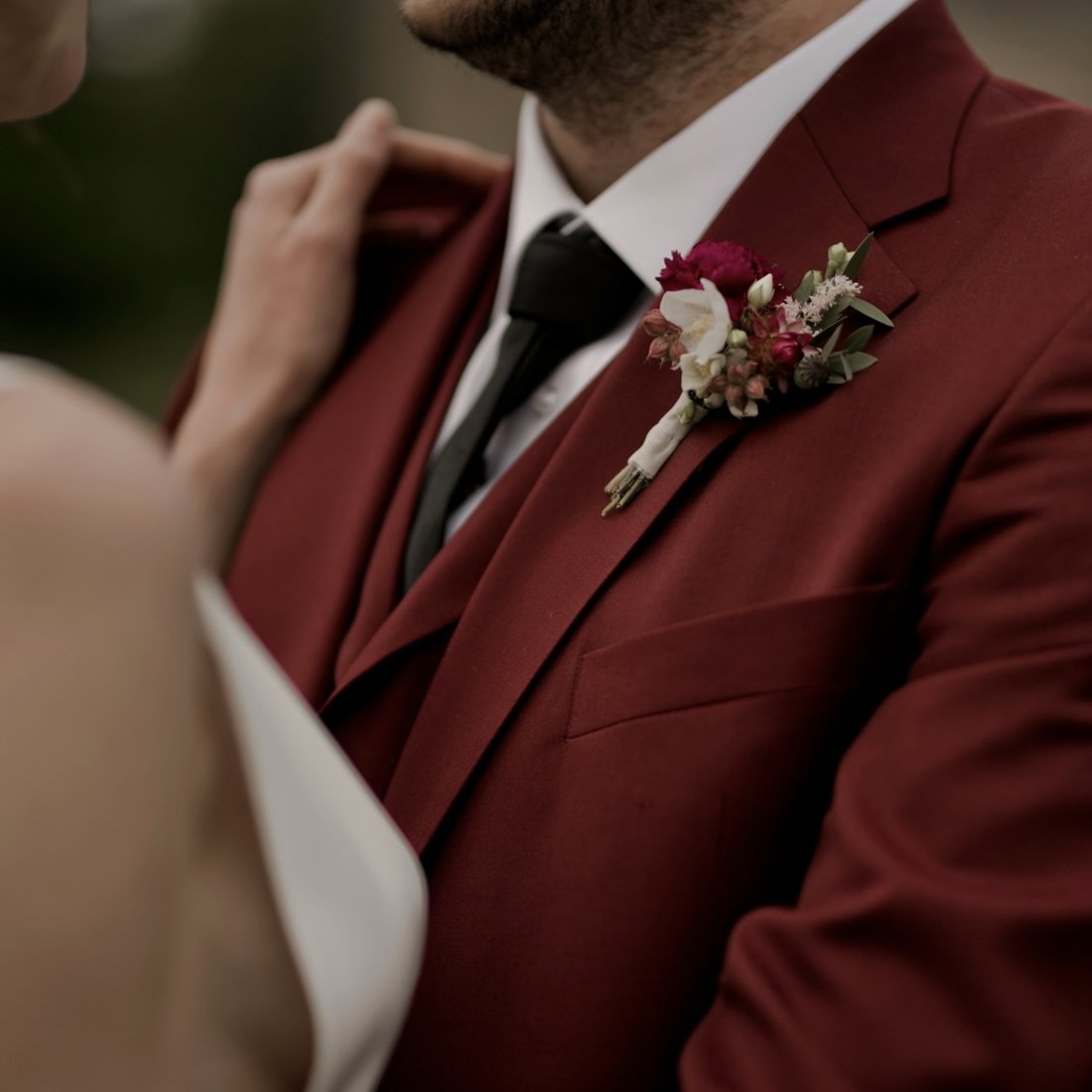 A deep burgundy three-piece wedding suit with a fresh buttonhole, the bride at his side