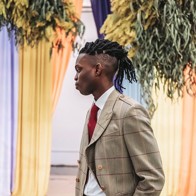 A double-breasted check suit with a deep red tie, worn amid hanging greenery and coloured drapes