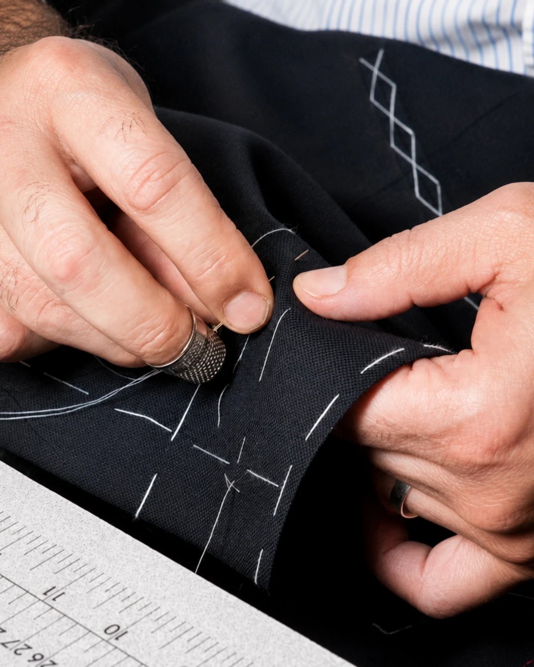 Hands with a thimble basting white tacking stitches into navy suiting