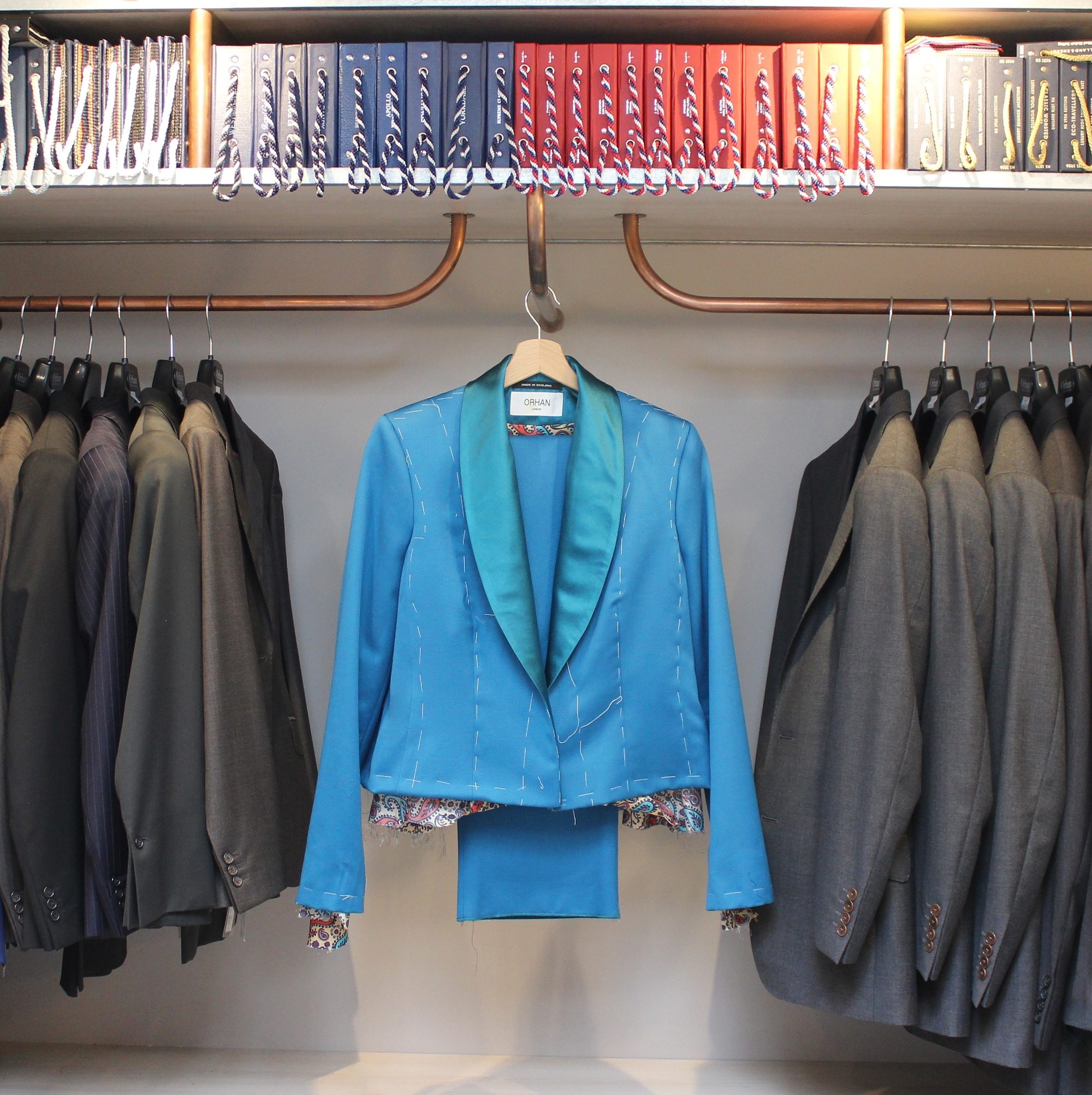 A basted turquoise jacket hanging on the studio rail among finished suits
